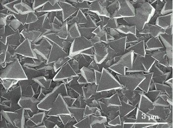 「図1.気相成長ダイヤモンド薄膜の電子顕微鏡写真　表面に三角形に成長した特徴的な結晶粒が見られるが、これは、{111}方向に成長したダイヤモンドである。良好な超伝導状態を得るためには、{111}方向に成長することが望ましい。」の画像